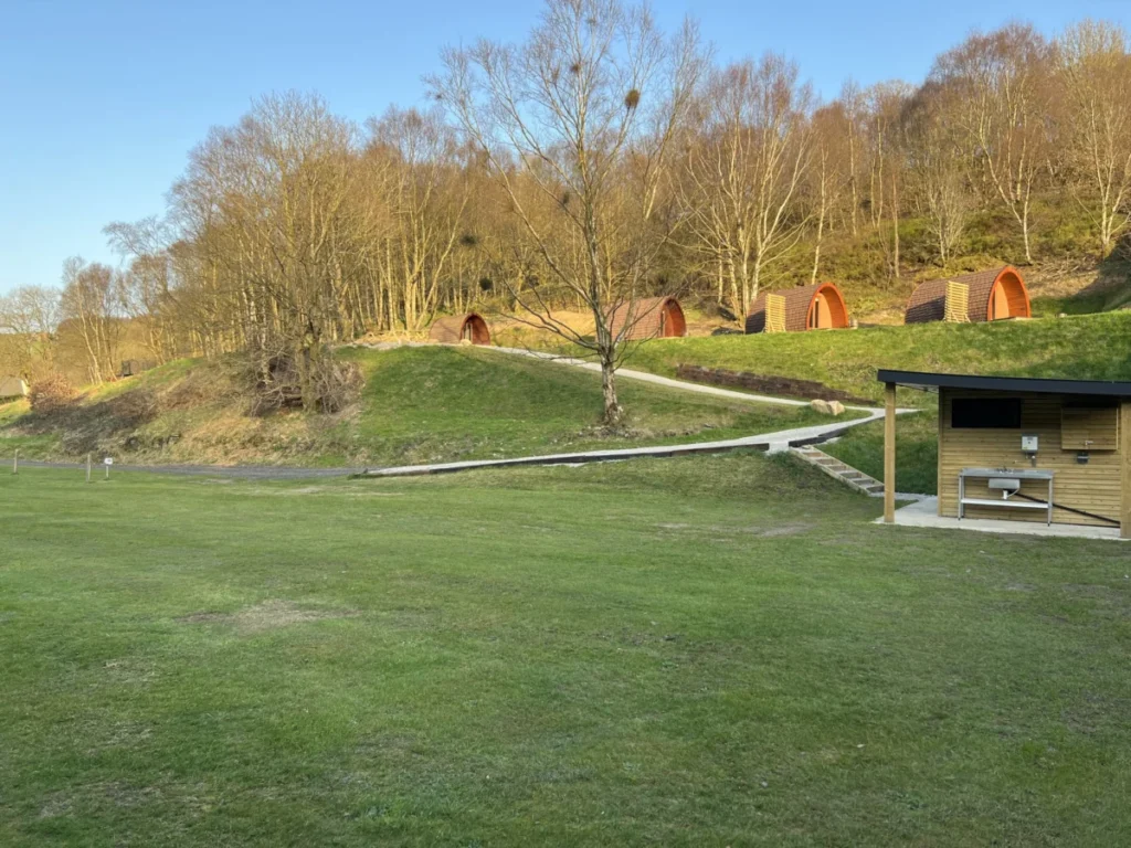 The camping field at Riverside Ripponden at golden hour, with the glamping pods on the bank above